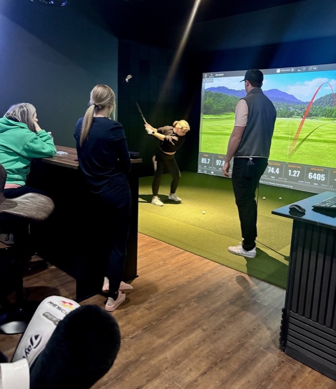 A golfer mid-swing in a FAIRWAYLAB bay while the coach watches her ball flight on the TrackMan screen