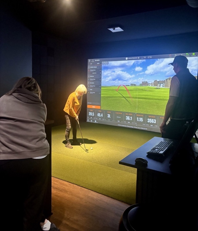 A golfer chipping with a wedge as the coach looks on, her shot numbers on the TrackMan screen